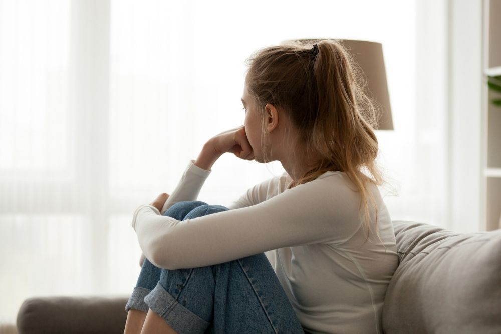 Young woman sitting on a couch with her knees up, looking out the window with a thoughtful expression