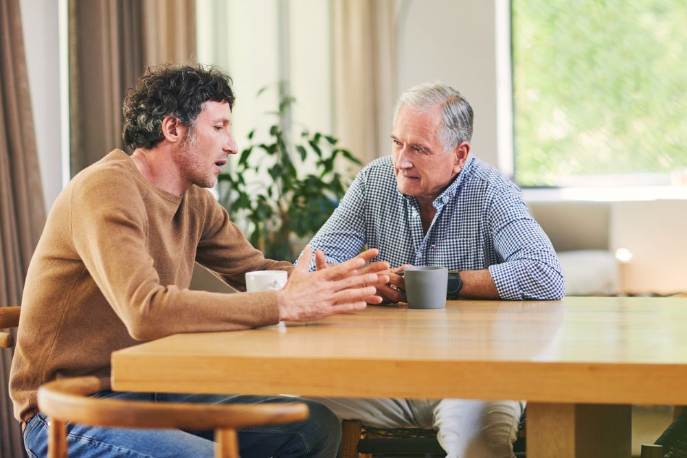 Two men sit at a wooden table having a serious conversation. The younger man gestures with his hands while the older man listens attentively. Both have coffee mugs in front of them.