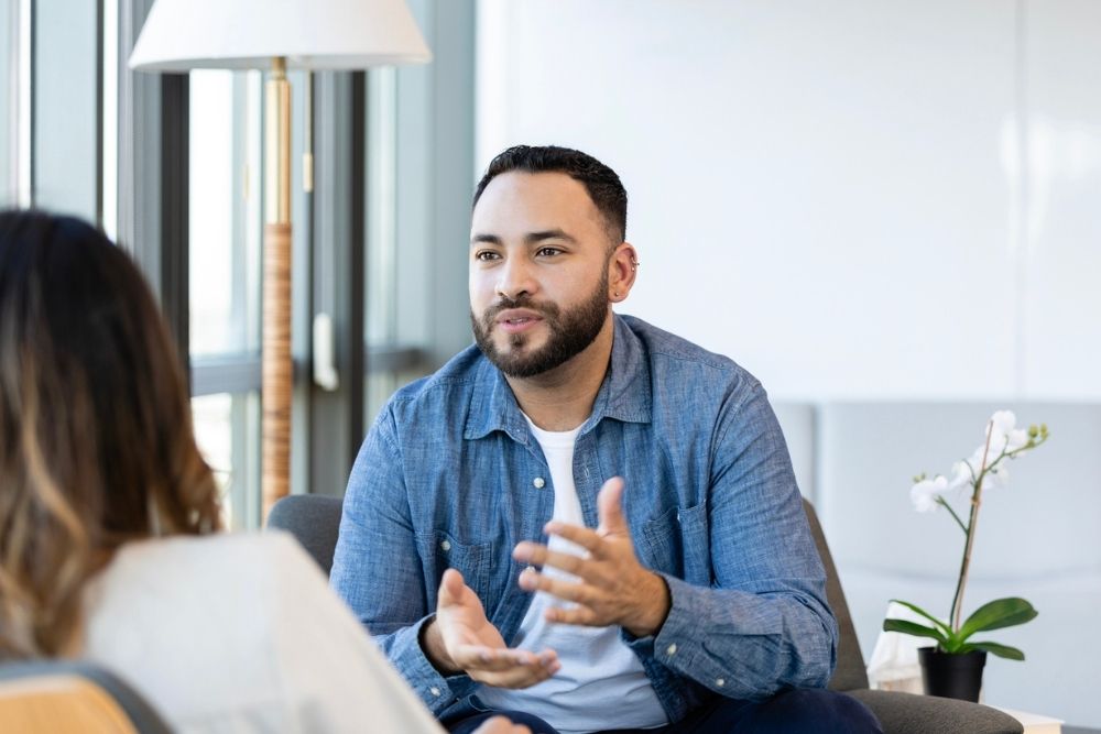 CBT (1000 x 667 px) A man in a casual blue shirt speaks while gesturing with his hands during a conversation with another person in a bright, modern office setting.