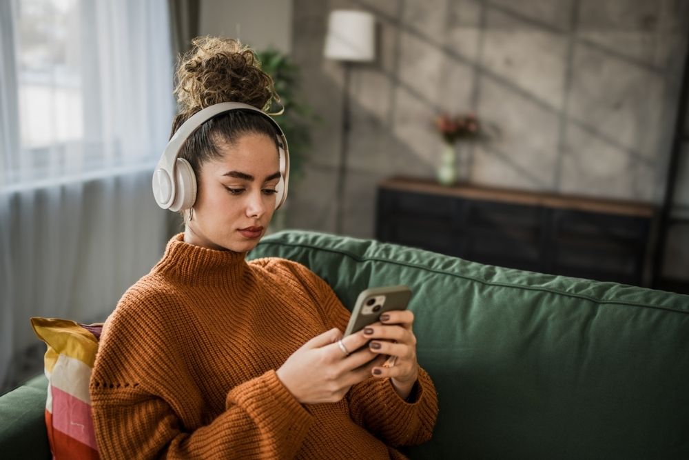 A young woman wearing headphones sits on a green couch, looking at her smartphone, appearing focused while listening to a podcast or music.