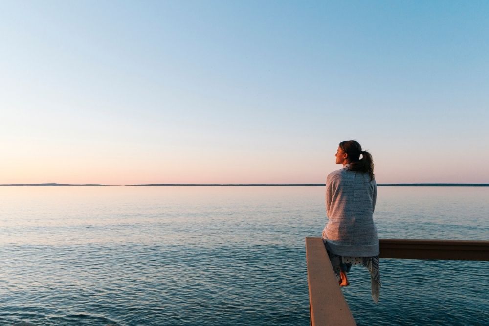 Woman sitting on a wooden railing by a calm lake at sunset, looking out over the water, symbolizing peace, reflection, and mindfulness.