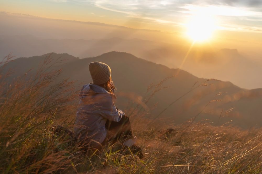 Sense of Purpose (1000 x 667 px) Person sitting on a grassy hilltop at sunrise, looking out over mountains and valleys, reflecting quietly in nature.