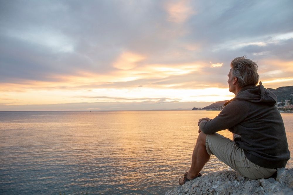A man sits on a rocky ledge overlooking the ocean at sunset, gazing thoughtfully at the horizon.