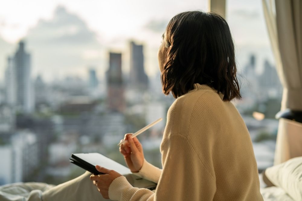 A person sits by a window overlooking a city skyline, holding a notebook and pen while reflecting quietly in a calm, sunlit room.