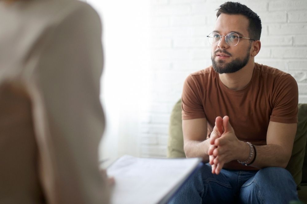 A man sitting and speaking with a therapist, hands clasped as he listens and engages in the conversation.
