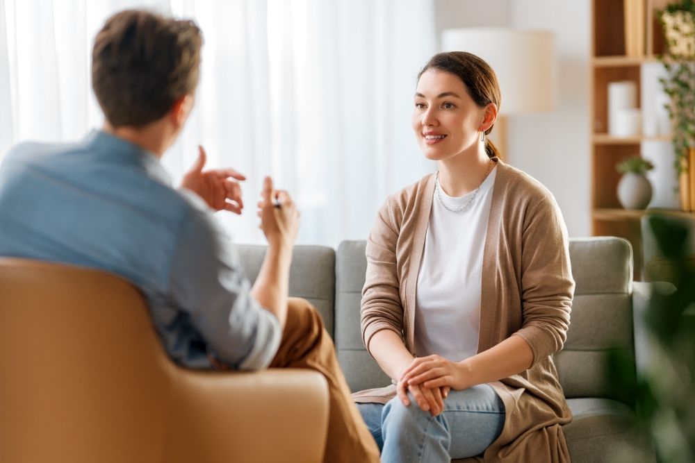 A woman sits on a sofa, smiling and engaged in conversation with a therapist discussion dual diagnosis treatment. The room is bright and warmly lit, conveying a relaxed, friendly atmosphere.